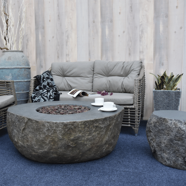 Elementi Boulder Rock Fire Pit Table OFG110 Without Flame, Coffee Cups, Propane Tank Cover and Accent Chairs and Couch on Background