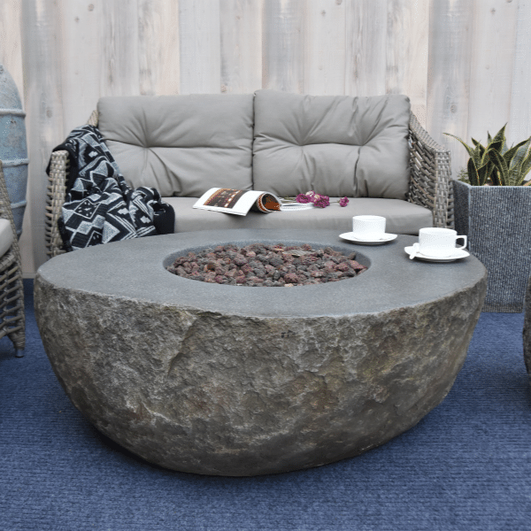 Elementi Boulder Rock Fire Pit Table OFG110 With No Flame, Coffee Cups and Accent Chairs and Couch on Background