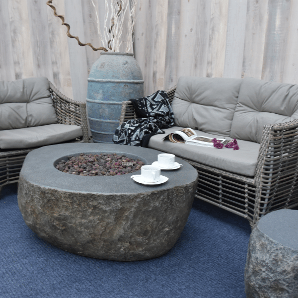 Elementi Boulder Rock Fire Pit Table OFG110 With No Flame, Coffee Cups, Matching Tank Cover, and Accent Chairs and Couch on Background